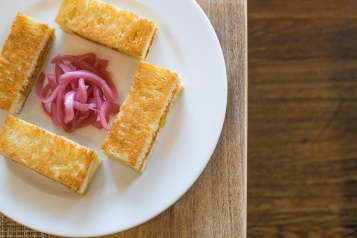 Three small rectangular sandwiches edge a white plate with pickled onion at its center.